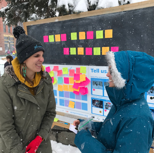 homeimage Two women smiling in front of an outdoor bulletin board with colourful notes on it